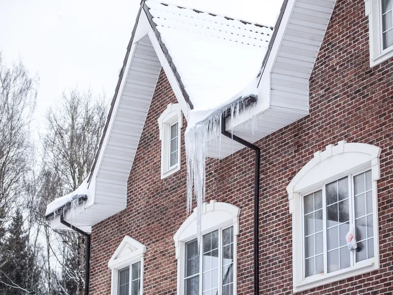 A thick ridge of ice and long icicles along the snowy eaves of a house, with a worker's steam wand melting a channel through the ice above the eavestrough.