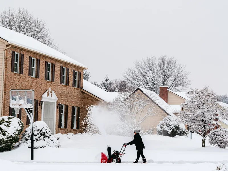 A person in a winter coat and toque clears fresh snow with a red walk-behind snow blower in front of a two-storey brick house with black shutters.